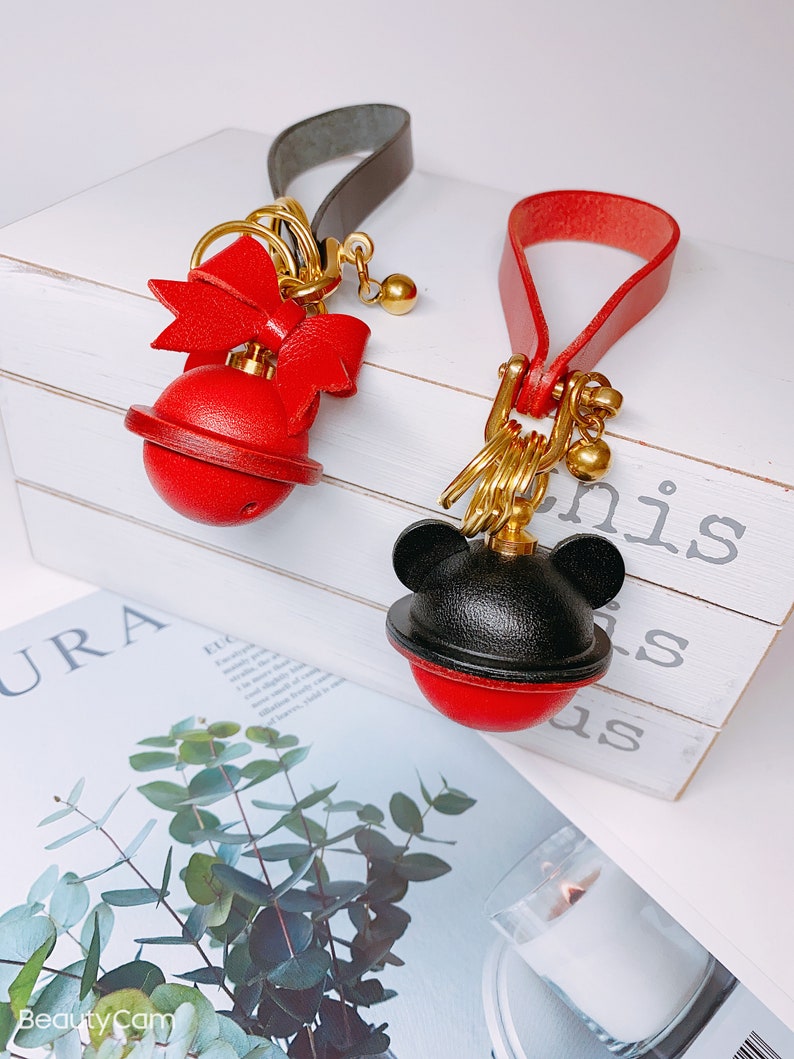 May include: Two red leather keychains with gold accents. One keychain has a bow and the other has a black Mickey Mouse head. Both keychains have a red bell with a gold ring.