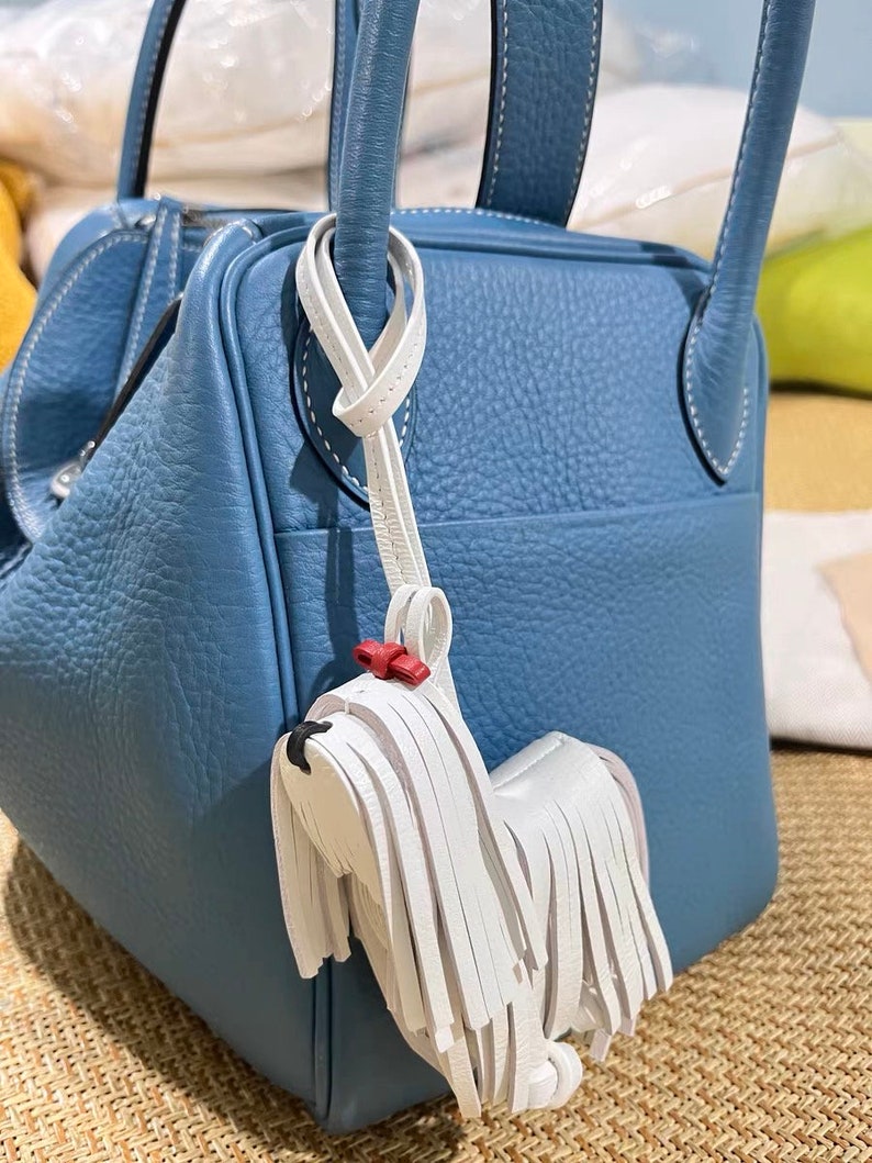 May include: A blue leather handbag with a white leather dog charm. The dog charm has a red bow and white fringe.
