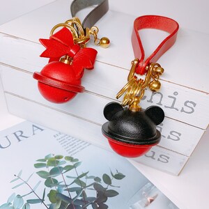 May include: Two red leather keychains with gold accents. One keychain has a bow and the other has a black Mickey Mouse head. Both keychains have a red bell with a gold ring.