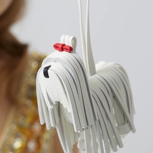 May include: White leather dog keychain with a red bow. The dog is made of strips of leather that resemble fur.
