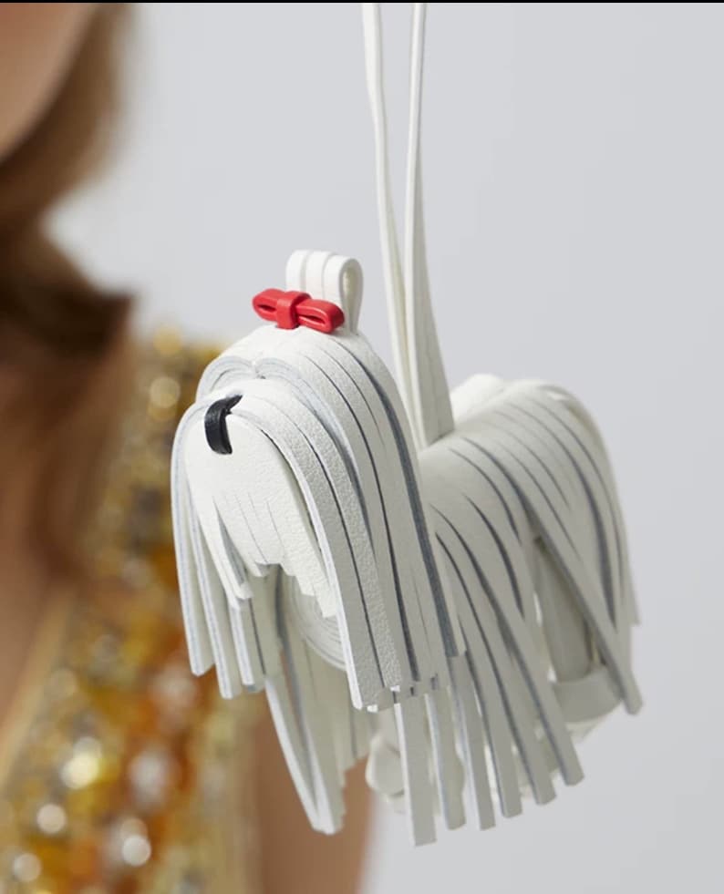 May include: White leather dog keychain with a red bow. The dog is made of strips of leather that resemble fur.