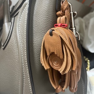 May include: A brown leather tassel keychain with a red bow attached to a silver hook. The keychain is hanging from a gray leather handbag.