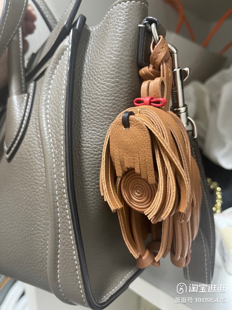 May include: A brown leather tassel keychain with a red bow attached to a silver hook. The keychain is hanging from a gray leather handbag.