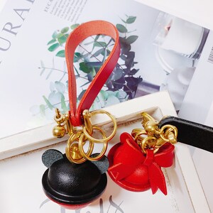 May include: Two leather keychains with gold key rings. One keychain features a black and red Mickey Mouse head with ears. The other keychain features a red hat with a bow. Both keychains are attached to red and black leather straps.