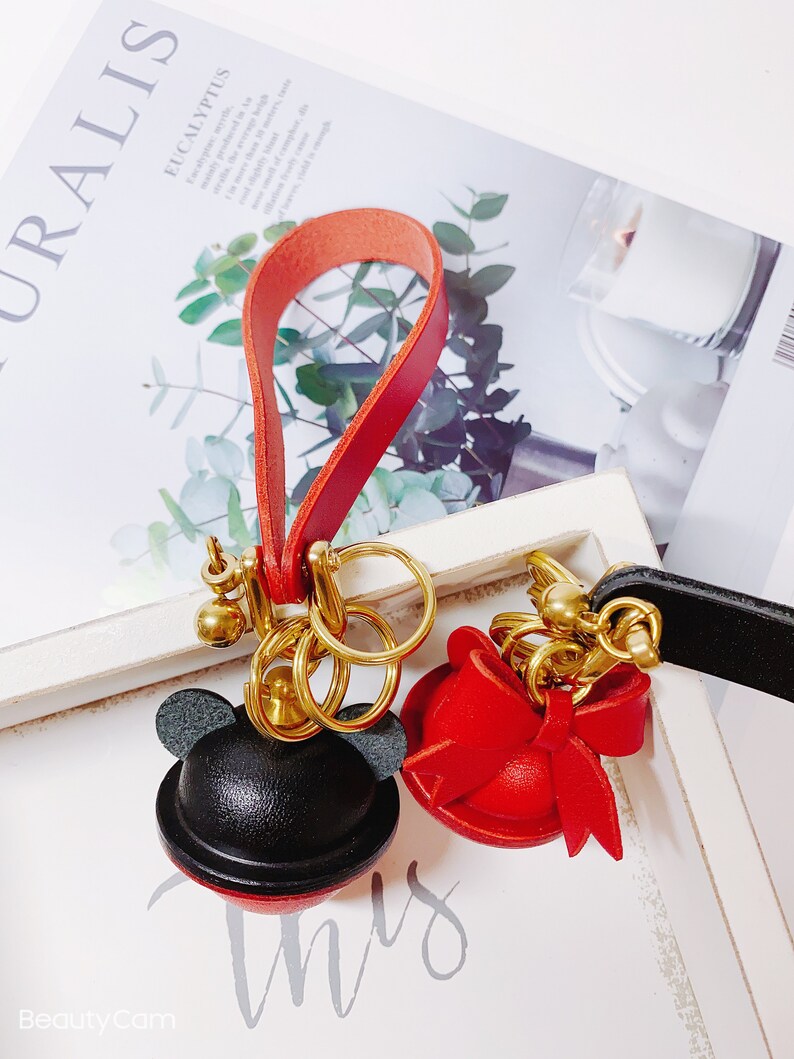 May include: Two leather keychains with gold key rings. One keychain features a black and red Mickey Mouse head with ears. The other keychain features a red hat with a bow. Both keychains are attached to red and black leather straps.