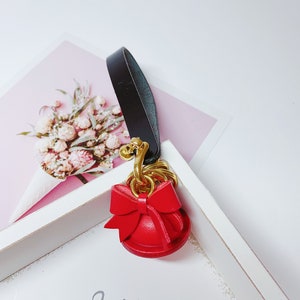 May include: A red leather keychain with a gold bell and a bow. The keychain is attached to a black leather strap.