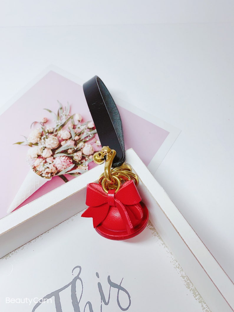 May include: A red leather keychain with a gold bell and a bow. The keychain is attached to a black leather strap.