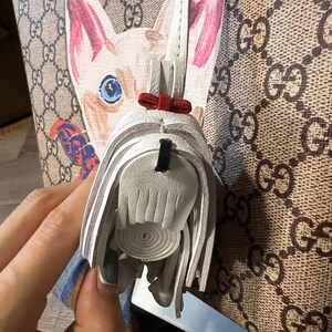 May include: A white leather purse charm with a fringe detail and a red bow. The charm is attached to a brown and beige patterned handbag.