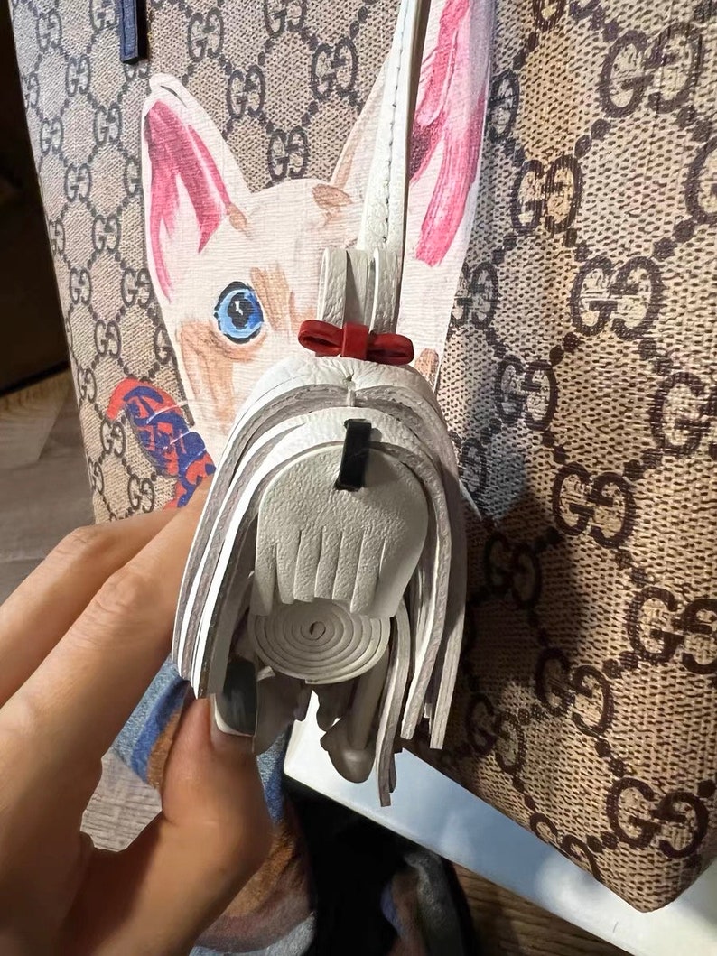 May include: A white leather purse charm with a fringe detail and a red bow. The charm is attached to a brown and beige patterned handbag.