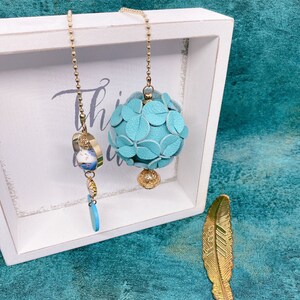 May include: Two blue leather tassel charms hanging from gold chains. The charms are in the shape of a ball and a heart. The charms are hanging from a white frame.