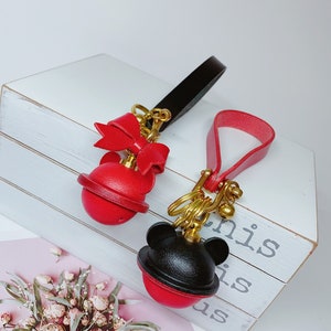 May include: Two leather keychains with red and black round charms. One charm has a red bow and the other has black mouse ears. Both charms have a gold metal ring and a gold metal bell.