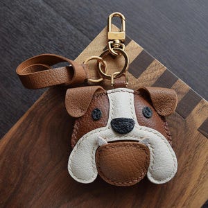 May include: A brown and white leather bulldog keychain with a gold key ring. The keychain is made of high-quality leather and features a detailed design of a bulldog's face.