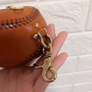 以下が含まれることがあります： ゴールドの金属製の留め具が付いた、野球ボールの形をした茶色の革製のハンドバッグです。