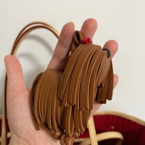 May include: A brown leather dog keychain with a red bow. The keychain is shaped like a Yorkshire Terrier and has a fringe of leather around the body.