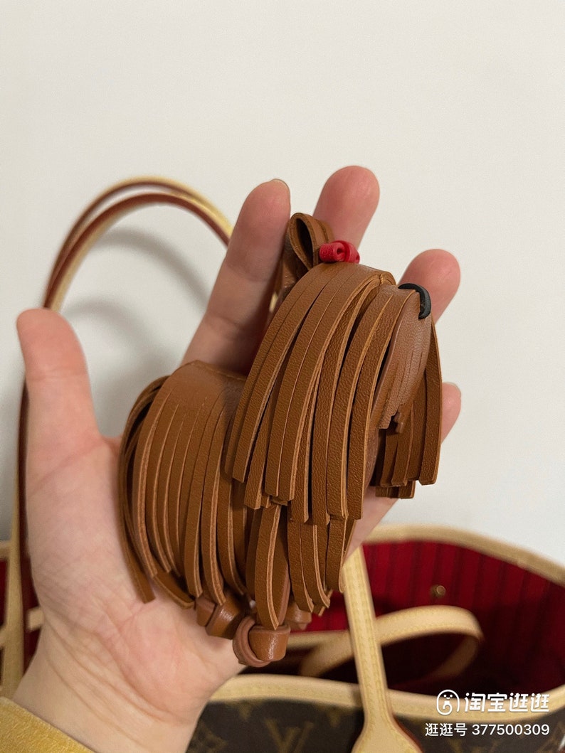 May include: A brown leather dog keychain with a red bow. The keychain is shaped like a Yorkshire Terrier and has a fringe of leather around the body.