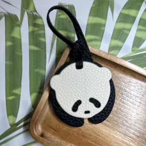 May include: A black and white panda-shaped leather keychain with a black loop. The keychain is sitting on a wooden tray.