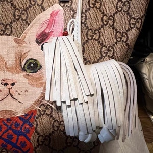 May include: A white leather tassel with a knotted end attached to a brown and beige handbag with a white cat print. The handbag has a repeating pattern of the letters "GG" throughout.