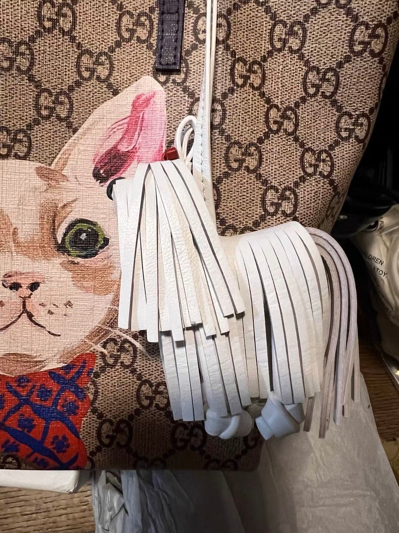 May include: A white leather tassel with a knotted end attached to a brown and beige handbag with a white cat print. The handbag has a repeating pattern of the letters "GG" throughout.