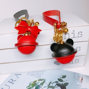 May include: Two red and black leather keychains with gold accents. One keychain features a red bow and the other has black mouse ears. Both keychains have a red ball with a gold bell inside.