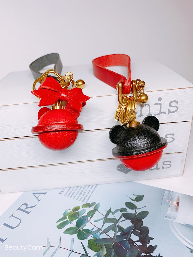 May include: Two red and black leather keychains with gold accents. One keychain features a red bow and the other has black mouse ears. Both keychains have a red ball with a gold bell inside.