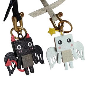 May include: Two keychains, one with a black devil character and the other with a white angel character. Both characters have wings and cute faces.