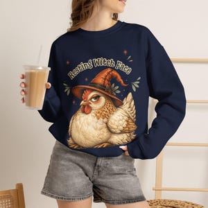 May include: A navy blue long-sleeve sweatshirt featuring a graphic of a chicken wearing an orange witch hat. The text above the chicken reads "Resting Witch Face" in a stylized font. The design includes small star and leaf accents in orange and green.