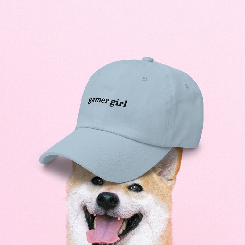 May include: A light blue baseball cap with the text "gamer girl" embroidered on the front. The cap is worn by a Shiba Inu dog.
