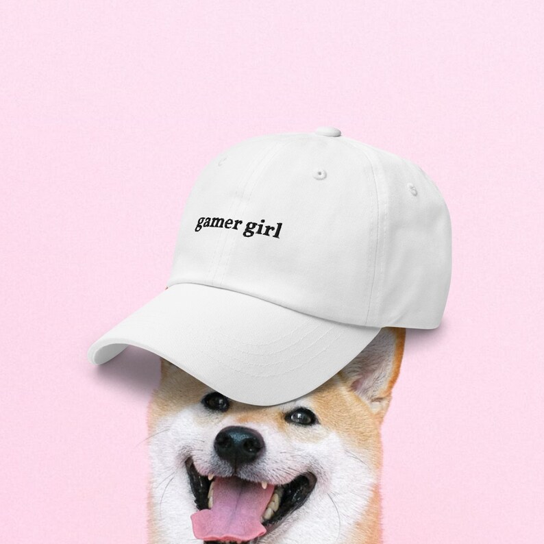May include: A white baseball cap with the text "gamer girl" embroidered in black on the front. The cap is worn by a Shiba Inu dog.
