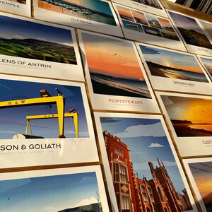 Puede incluir: Una colecci&oacute;n de impresiones ilustradas coloridas que representan varios monumentos y escenas de Irlanda del Norte. Las impresiones se muestran en una superficie de madera e incluyen lugares como Glens of Antrim, Samson & Goliath, Portstewart, Brown's Bay y Castlerock.