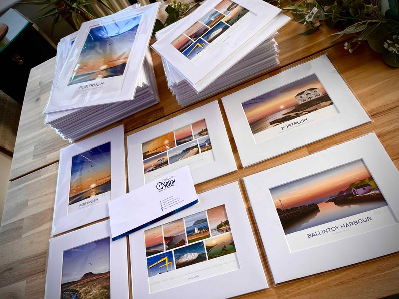 Puede incluir: Una colecci&oacute;n de impresiones enmarcadas que muestran paisajes pintorescos de la costa de Irlanda del Norte. Las impresiones se muestran en una mesa de madera e incluyen im&aacute;genes de Porttrush y Ballintoy Harbour. Las impresiones est&aacute;n todas en marcos blancos con un passe-partout blanco.