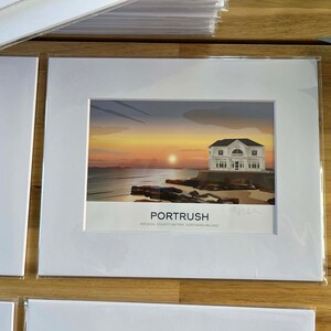 Puede incluir: Una impresi&oacute;n enmarcada en blanco de una puesta de sol sobre el oc&eacute;ano con una casa blanca en la costa. La impresi&oacute;n dice "Portrush, Arcadia, County Antrim, Northern Ireland" en la parte inferior.