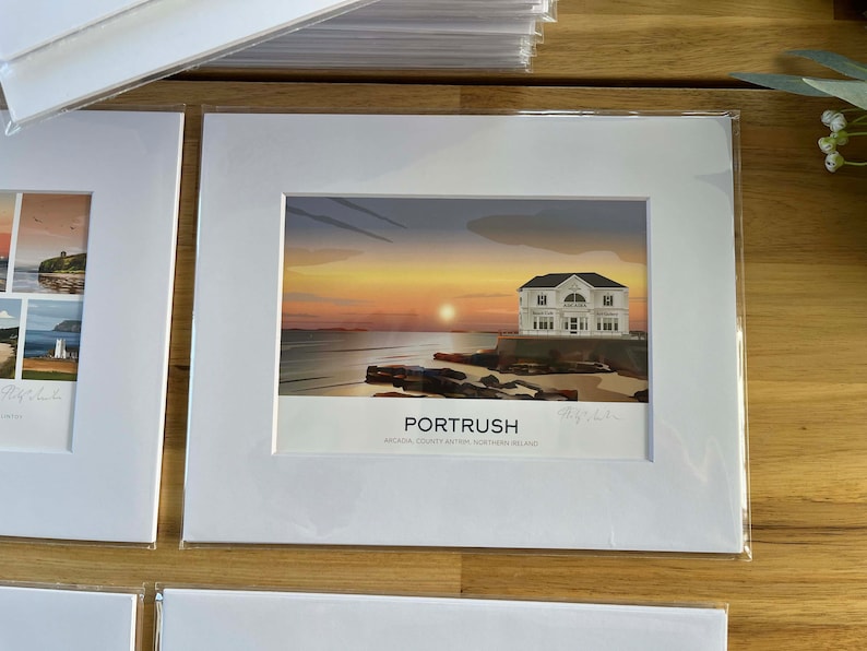 Puede incluir: Una impresi&oacute;n enmarcada en blanco de una puesta de sol sobre el oc&eacute;ano con una casa blanca en la costa. La impresi&oacute;n dice "Portrush, Arcadia, County Antrim, Northern Ireland" en la parte inferior.