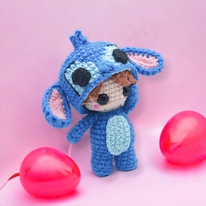 Puede incluir: Un juguete de peluche azul tejido a crochet que lleva un disfraz de Stitch azul y rosa. El juguete tiene el pelo castaño y los ojos negros. El juguete está de pie sobre una superficie rosa con dos globos rosas.