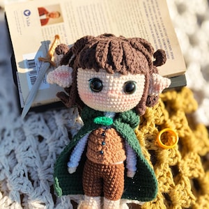 Könnte beinhalten: Eine handgefertigte Häkelpuppe mit braunen Haaren, einem grünen Umhang und braunen Hosen. Die Puppe hat große schwarze Augen und hält ein kleines Schwert. Ein Buch und ein goldener Ring sind ebenfalls im Bild.