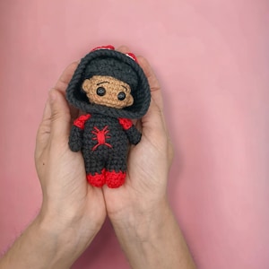 Puede incluir: Muñeca de ganchillo hecha a mano con un disfraz de superhéroe. La muñeca lleva un mono con capucha gris oscuro con detalles rojos en los brazos y los pies. Un emblema de araña roja está en el pecho. La muñeca se sostiene con dos manos sobre un fondo rosa.