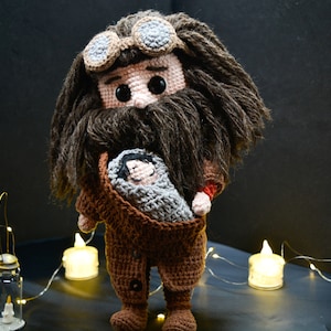 Puede incluir: Un juguete de peluche de crochet de un mago sosteniendo un bebé con un atuendo marrón y gris. El mago tiene una barba marrón y lleva gafas redondas.