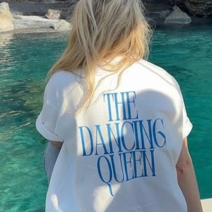 Puede incluir: Camiseta blanca con el texto azul "THE DANCING QUEEN". La persona está de espaldas a la cámara, con el pelo rubio. El fondo muestra una masa de agua y formaciones rocosas.