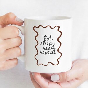 Puede incluir: Taza de cerámica blanca con las palabras "Eat sleep read repeat" en un marco ondulado marrón. La taza está sujeta por una persona de piel clara. La taza tiene un asa en el lado izquierdo.