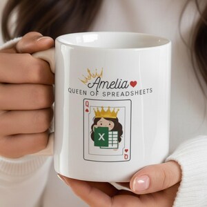 Puede incluir: Taza de cerámica blanca con el texto "Amelia Queen of Spreadsheets", un diseño de corona y corazón. La taza también muestra una carta de juego con una mujer de dibujos animados con una corona y una hoja de cálculo de Excel.