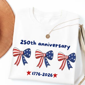 Puede incluir: Camiseta blanca con un diseño patriótico. La camiseta presenta tres ilustraciones de pajaritas con rayas rojas y blancas y patrones de estrellas azules. El texto "250th anniversary" y "1776-2026" también está impreso.