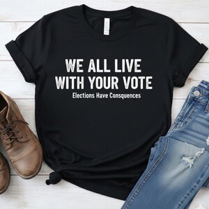 Puede incluir: Camiseta negra con el texto blanco "WE ALL LIVE WITH YOUR VOTE" y "Elections Have Consequences". La camiseta se combina con vaqueros azules y zapatos de cuero marrón. La camiseta está anudada en la parte inferior.