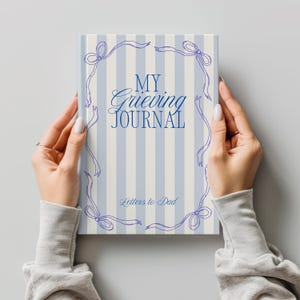 Puede incluir: Un diario titulado "My Grieving Journal" con el subtítulo "Letters to Dad". La portada presenta un patrón de rayas azul claro y blanco con un borde decorativo de lazos y cintas. El diario está sostenido por dos manos.