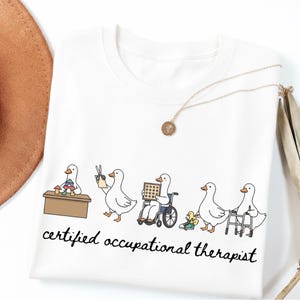 Peut inclure: T-shirt blanc avec un motif de canards dans des situations de thérapie occupationnelle. Le texte "certified occupational therapist" est imprimé en noir. Un collier doré avec un pendentif est posé sur le t-shirt.