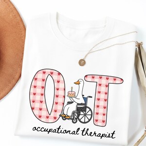 Peut inclure: T-shirt blanc avec un motif graphique représentant les lettres "OT" en vichy rose avec des cœurs rouges. Un canard de dessin animé en fauteuil roulant tient un gâteau. Les mots "occupational therapist" sont imprimés en dessous.