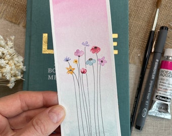 May include: Watercolor bookmark featuring a gradient of pink and blue hues. Delicate floral stems with colorful blooms are painted on the lower half. The bookmark is held by a hand, with art supplies and a book in the background.