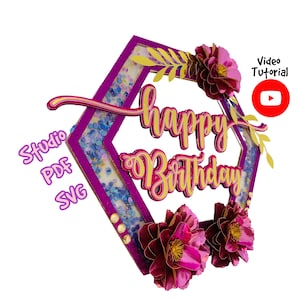 May include: A purple hexagonal frame with glitter and gold accents, featuring the words "Happy Birthday" in pink and gold lettering. The frame is decorated with three pink paper flowers.