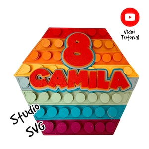 May include: A colorful hexagonal birthday card with a rainbow pattern and a number 8 in the center. The name "CAMILA" is written in a bold font below the number. The card is made to look like a Lego brick.