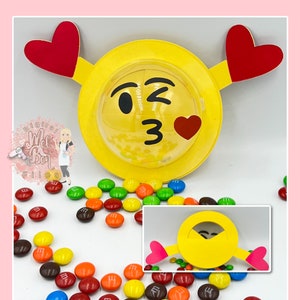 Pode incluir: Um emoji amarelo com um olho piscando e um beijo soprado, com um coração vermelho no centro do emoji. O emoji está preso a um círculo amarelo com dois corações vermelhos nas laterais. O emoji está rodeado de doces M&M's coloridos.