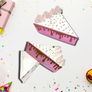 Puede incluir: Dos cajas de cartón en forma de porción de pastel rosa y blanca con un diseño de glaseado de chocolate y chispas de colores. Las cajas están abiertas y listas para llenarse de golosinas.