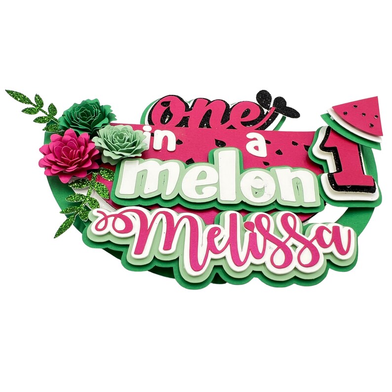 May include: One in a melon cake topper with green leaves, pink and green flowers, and the name Melissa. The topper is decorated with glitter and has a watermelon slice design.